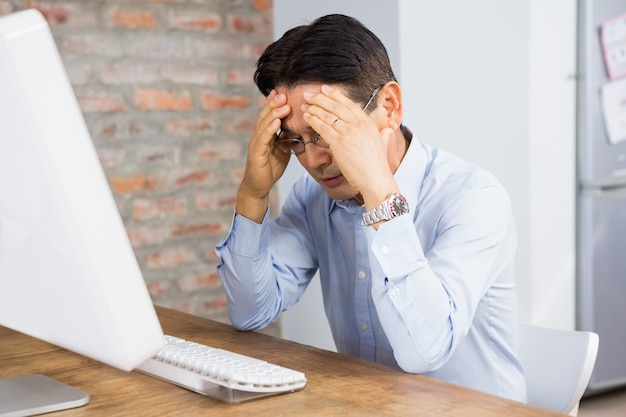 Leidender Mann mit den Händen auf dem Gesicht, das am Schreibtisch mit Computer auf ihm sitzt