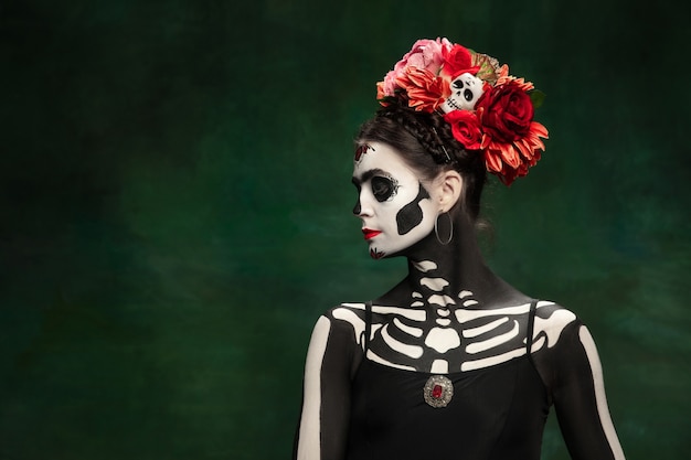 Leidenschaftlich. Junges Mädchen wie Santa Muerte Saint Death oder Sugar Skull mit hellem Make-up. Porträt lokalisiert auf dunkelgrünem Studiohintergrund mit Exemplar. Feiern von Halloween oder Tag der Toten.