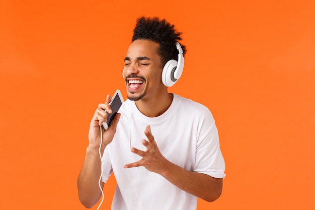 Leidenschaftlicher und sorgloser Afroamerikaner-Typ, der träumt, wird echter Sänger, hält Telefon und singt in Handy, während er Karaoke-App spielt und Kopfhörer trägt, orange Wand