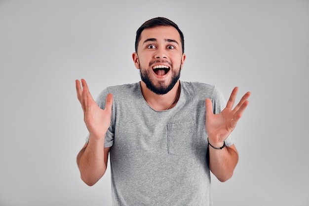 Leistung, Erfolgskonzept. Ein fröhlicher Kaukasier hebt die Hände, seine Augen sind voller Glück und freuen sich über seine großen Leistungen. Attraktiver schwarzer Mann in einem grauen T-Shirt.