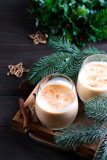 Leite de gemada Natal coquetel com canela, servido em dois copos na bandeja vintage com ramo de abeto no escuro de madeira