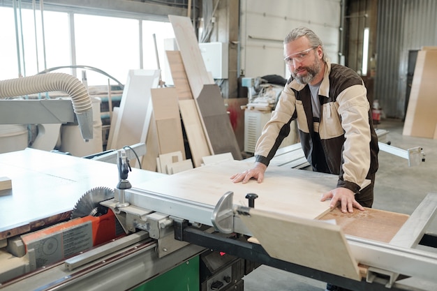 Leitender Arbeiter der Möbelherstellung, der eine rechteckige Platte auf der Werkbank befestigt, bevor er sie mit einem elektrischen Handwerkzeug schneidet oder schleift