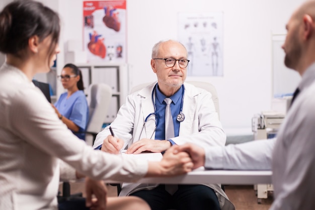 Leitender Arzt, der dem jungen Paar während der medizinischen Untersuchung im Krankenhausbüro schlechte Nachrichten gibt. Mann und Frau, die Händchen halten. Krankenschwester, die Patientenröntgenaufnahme betrachtet.