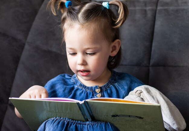 Leitura infantil. Menina bonita 1-3 está lendo um livro com uma lebre de pelúcia de brinquedo sentada em um sofá.