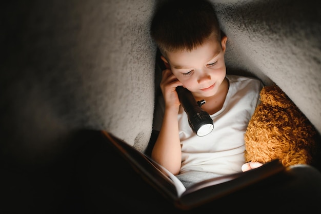 Lendo livro e usando lanterna menino em roupas casuais, deitado perto da tenda à noite