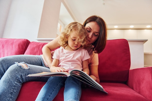 Lendo um grande livro Jovem mãe com sua filha em roupas casuais juntos dentro de casa em casa
