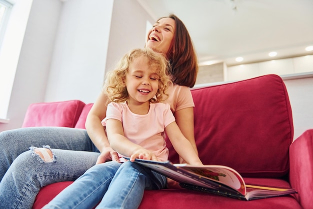 Lendo um grande livro Jovem mãe com sua filha em roupas casuais juntos dentro de casa em casa