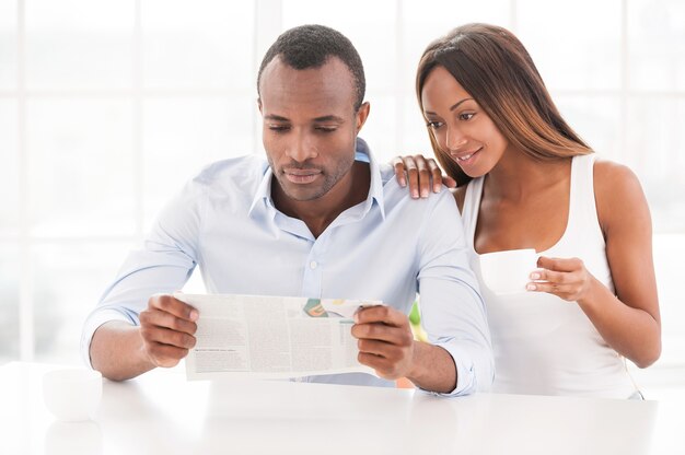 Lendo um novo jornal juntos. Lindo casal jovem africano sentado perto um do outro lendo jornal, enquanto uma mulher segurando uma xícara de café e sorrindo