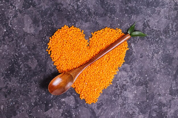 Lentejas de naranja cruda para una nutrición y salud adecuadas en forma de corazón con una cuchara de madera y hojas sobre un fondo gris