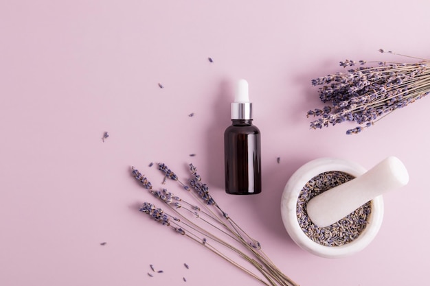 Óleo cosmético de lavanda em uma garrafa com pipeta de vidro em um fundo roxo com galhos de plantas e sementes em uma vista superior de argamassa