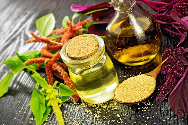 Óleo de amaranto em uma jarra de vidro e sementes de amaranto decantador em uma colher flores verdes e roxas marrons e folhas de plantas no fundo da placa de madeira preta