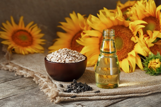 Óleo de girassol em uma garrafa de vidro com sementes em uma xícara e flores em uma mesa de madeira.