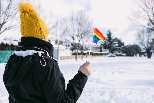 Lesbenfrau mit dem Rücken drehte sich mit gelber Schneekappe in einem schneebedeckten Gebiet, das lgbt Regenbogenfahne hält