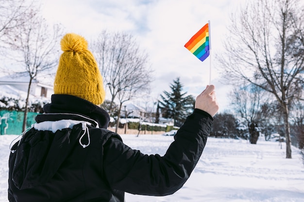 Lesbenfrau mit dem Rücken drehte sich mit gelber Schneekappe in einem schneebedeckten Gebiet, das lgbt Regenbogenfahne hält