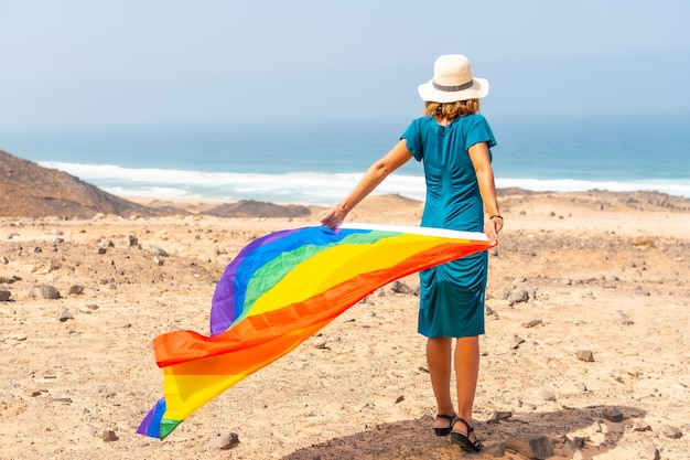 Una lesbiana con vestido verde, sombrero blanco y con la bandera LGBT junto al mar, símbolo de la homosexualidad.