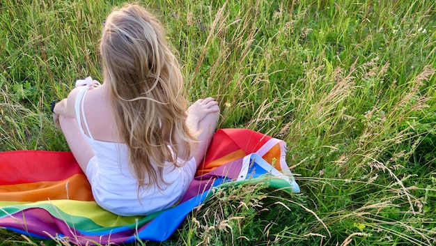 Lesbische bisexuelle blonde Frau sitzt auf LGBT-Flagge auf Gras in weißem Kleid Unterstützung der nicht-traditionellen Orientierung im Monat der Würde, dem Tag der Bisexualität