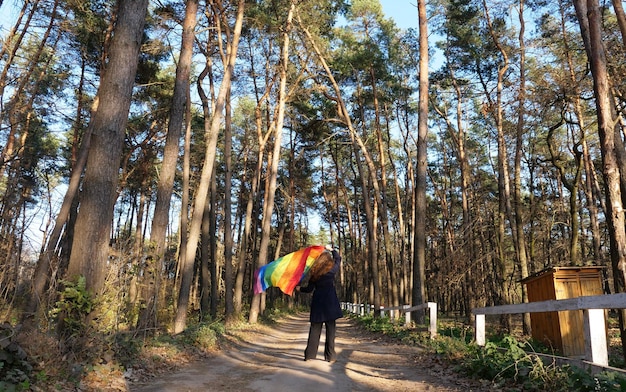 Lesbische bisexuelle Frau, die eine Regenbogen-LGBT-Flagge hält und im Monat des Stolzes auf dem Weg im Wald spazieren geht und eine Schwulenparade Bisexuality Day oder National Coming Out Day feiert