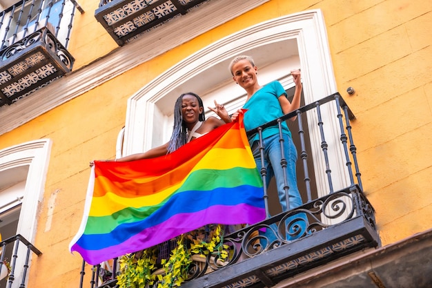 Lesbische Mädchenpaare mit LGBT-Flagge auf dem Balkon zu Hause feiern lächelnden LGBT-Stolz