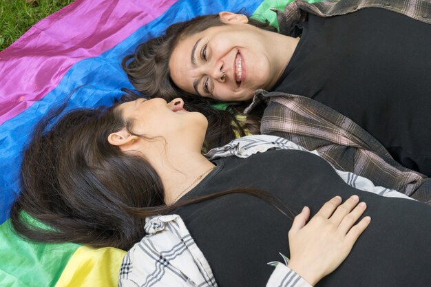 Lesbisches Paar trägt eine LGBT-Regenbogenflagge auf dem Boden liegender liebevoller Moment zwischen zwei Frauen ...