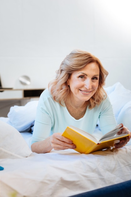 Lesen interessantes Buch. Ansprechende Frau mit breitem Lächeln, die ein gelbes Hardcover-Buch trägt, während sie auf einem unfertigen Bett liegt