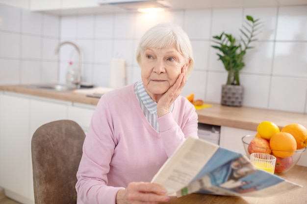 Lesen. Ältere grauhaarige Frau, die eine Zeitung in der Küche liest