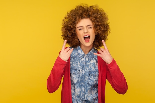 Let39s rock Retrato de una mujer loca con el pelo rizado en ropa informal que muestra el signo del rock and roll y ruge furiosamente luciendo agresivo y desobediente en un estudio interior aislado en un fondo amarillo