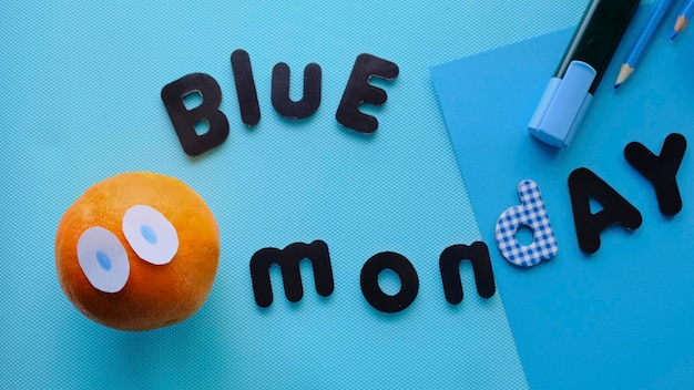 Letras azules del lunes sobre fondo azul con una naranja con ojos y papelería azul. alegre lunes