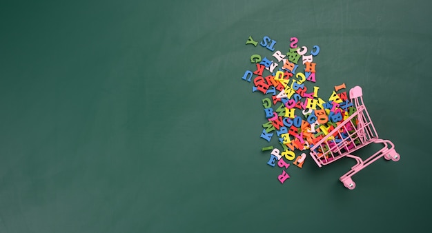 Letras multicoloridas do alfabeto inglês espalhadas em um carrinho de compras em miniatura em uma superfície verde, vista de cima, espaço para cópia