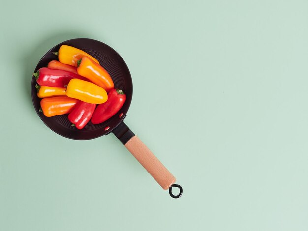 Leuchtend rote, orange und gelbe ganze frische kleine Paprikaschoten in einem Topf mit Holzgriff. Draufsicht isoliert auf einem grünen Hintergrund mit Kopierbereich