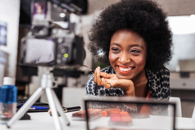 Leuchtenden. Lächelnde Afroamerikanerfrau in einem orangefarbenen Oberteil, das zur Kamera lächelt, während sie ein Make-up-Tutorial durchführt