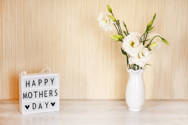 Leuchtkasten mit Schriftzug Glücklicher Muttertag, weiße Blumen Eustoma oder Lisianthus in Vase auf Holztisch mit Kopierraum.