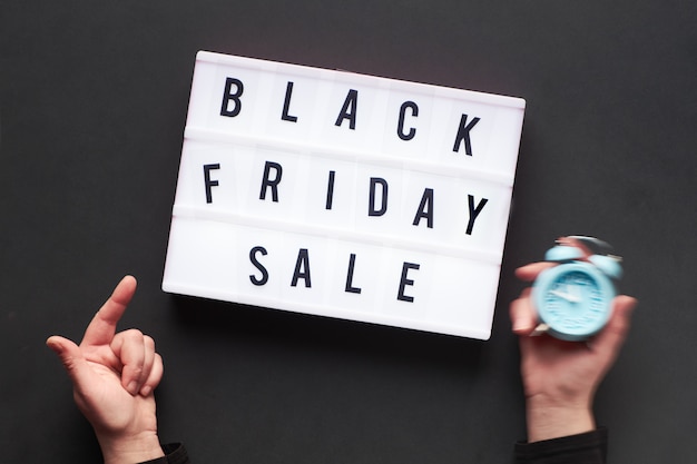 Leuchtkastentext "Black Friday Sale" mit zwei Zeigern, von denen einer den Wecker hält und der andere auf den Text im Leuchtkasten zeigt.