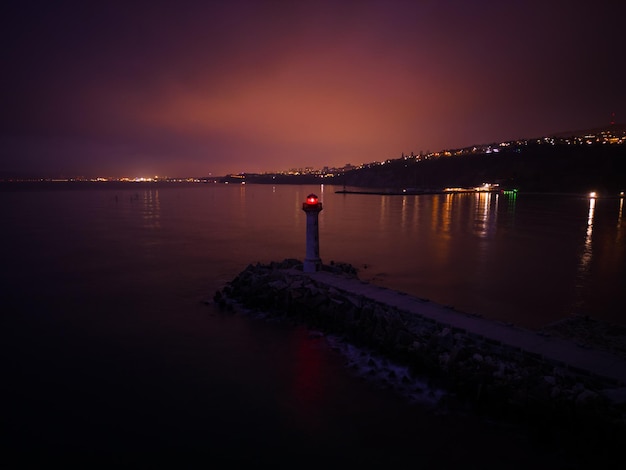 Leuchtturm an der Küste mit rotem Licht in der Nacht vor dem Hintergrund der Lichter der Schiffe