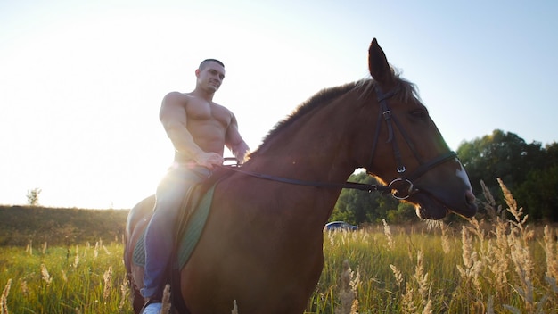 Levantador de pesas hombre sentado sobre un caballo que mastica hierba en medio del campo en el verano