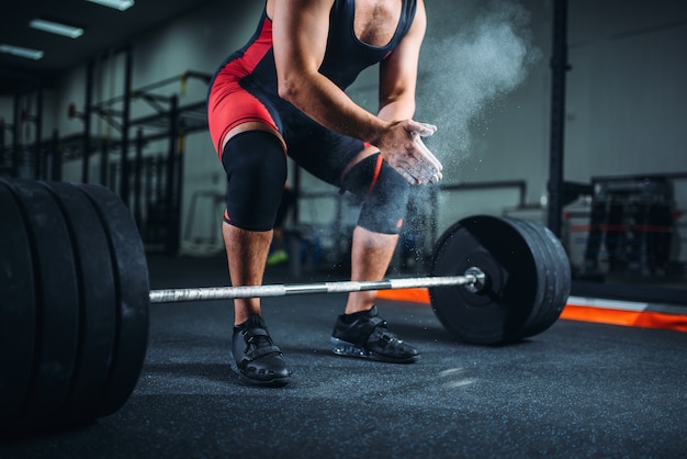 Levantador de pesas masculino en ropa deportiva se frota las manos con talco, preparación para el ejercicio con barra en el gimnasio.