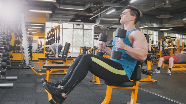 Levantamento de peso na academia - atleta musculoso realiza treinamento para bíceps com halteres, close-up tiro