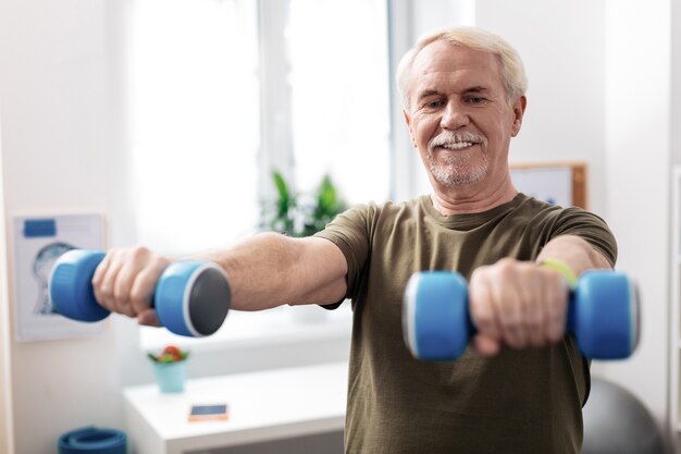 Levantando halteres. Homem simpático e alegre segurando as mãos à frente enquanto levanta halteres