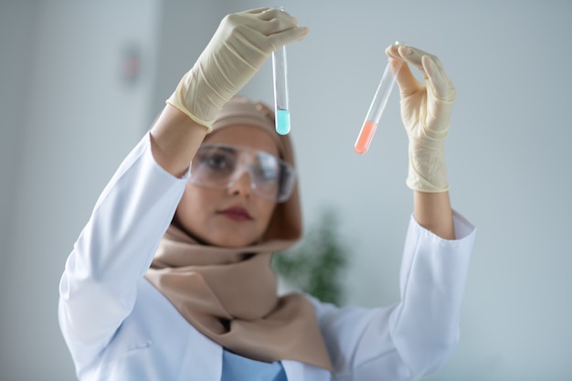 Levantando tubos de ensayo. Químico femenino vistiendo bata blanca levantando tubos de ensayo con productos químicos mientras los estudia