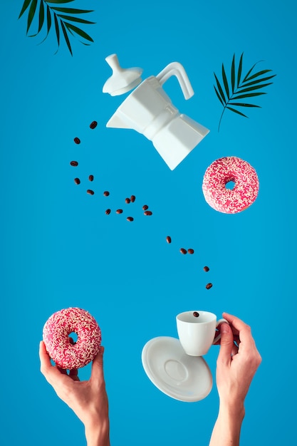 Levitação na moda. Linha voadora de grãos de café entre a cafeteira de cerâmica e a xícara de café expresso com Pires. Mãos femininas segurar rosquinhas rosa com açúcar granulado. Parede de hortelã azul com folhas de palmeira.
