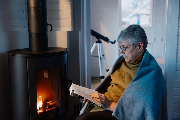 Foto leyendo un libro junto a la chimenea la abuela está sentada junto a la cálida chimenea con un libro en las manos y está leyendo cuidadosamente una historia emocionante
