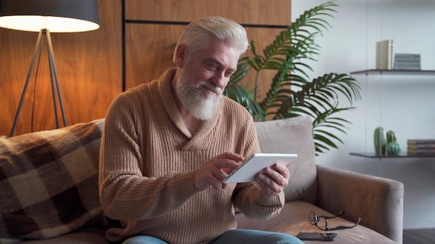 Leyendo noticias. Hombre senior con barba gris con tableta digital sentado en el sofá en casa