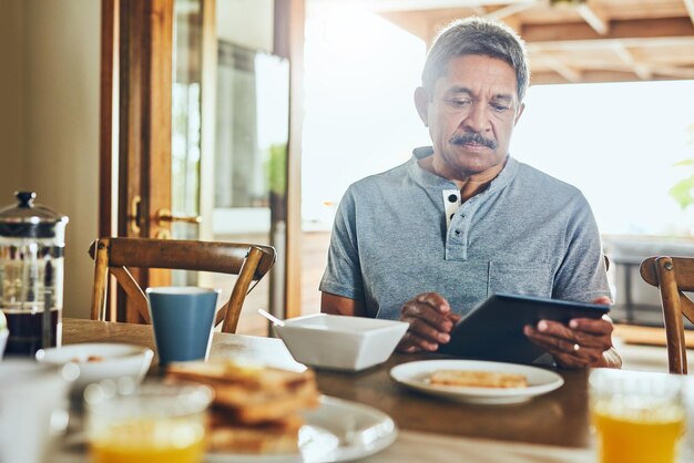 Leyendo las noticias de la manera moderna Foto de un anciano concentrado navegando en una tableta mientras desayuna una mesa en casa