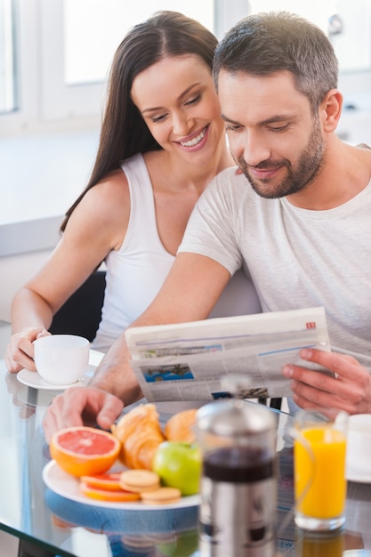 Leyendo periódicos nuevos juntos. Hermosa joven pareja uniéndose y leyendo el periódico juntos mientras está sentado en la cocina y desayunando