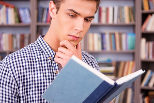 Leyendo su libro favorito. Hombre joven concentrado leyendo un libro y sosteniendo la mano en la barbilla mientras está de pie contra la estantería