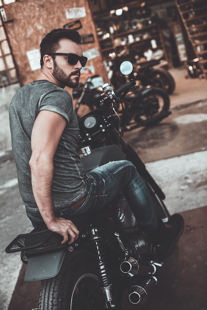 La libertad corre por sus venas. Vista trasera del joven confiado sentado en su bicicleta y mirando por encima del hombro con garaje para motocicletas en el fondo