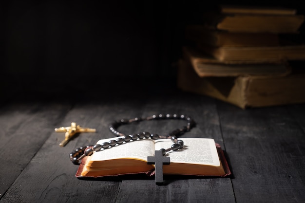 Libro abierto de la Biblia y crucifijo en mesa oscura. Imagen de bajo perfil del Nuevo Testamento, cruz y rosario en luz brillante entre oscuridad y sombras con espacio de copia