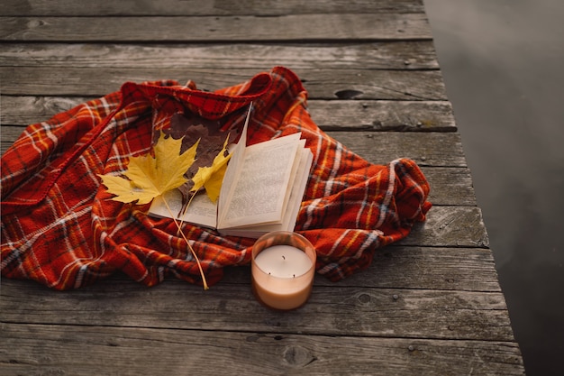 Libro abierto con vela y ramo de hojas de otoño con un plaid sobre un fondo de madera