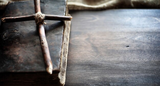 Libro antiguo religioso sobre una mesa de madera. Una cruz religiosa atada con una cuerda y arpillera junto a la Biblia. Adoración, pecados y oración.