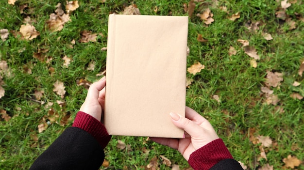 Un libro cerrado en una cubierta hecha de papel artesanal en manos femeninas con pasto verde y hojas amarillas caídas en el fondo. Vista plana endecha, superior. Plantilla, diseño. Copie el espacio.