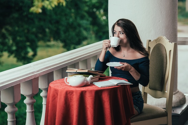 libro de lectura de la chica joven mientras que bebe el café en el día soleado que se sienta en el balcón.